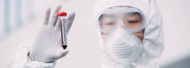 panoramic shot of asian epidemiologist in hazmat suit and respirator mask looking at test tube with blood sample while standing outdoors