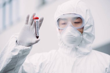 asian epidemiologist in hazmat suit and respirator mask looking at test tube with blood sample while standing outdoors