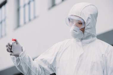 asian epidemiologist in hazmat suit and respirator mask holding test tube with blood sample while standing outdoors