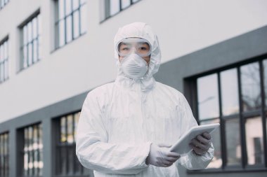 asian epidemiologist in hazmat suit and respirator mask holding digital tablet and looking at camera while standing on street