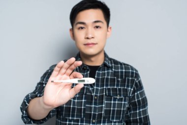 selective focus of young asian man holding thermometer while looking at camera on grey background