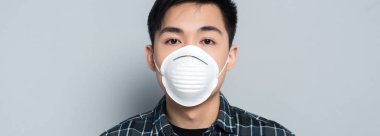 panoramic shot of young asian man in respirator mask looking at camera on grey background