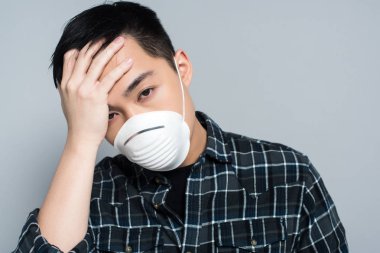 young asian man in respirator mask touching forehead and looking at camera while suffering from headache isolated on grey