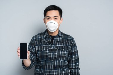 young asian man in respirator mask showing smartphone with blank screen and looking at camera on grey background