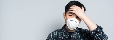 panoramic shot of young asian man in respirator mask touching forehead and looking at camera while suffering from headache isolated on grey