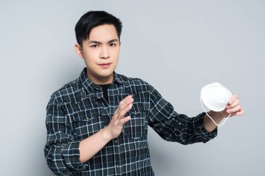 young asian man showing refuse gesture while holding respirator mask and looking at camera on grey background