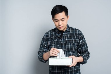 young asian man holding pack of paper napkins while suffering from runny nose on grey background clipart