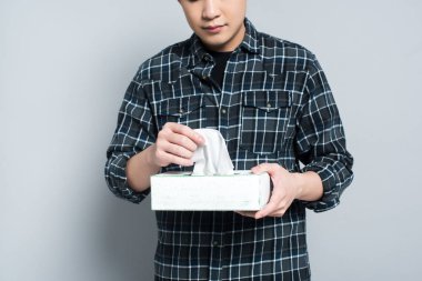 cropped view of young man holding pack of paper napkins while suffering from runny nose on grey background clipart