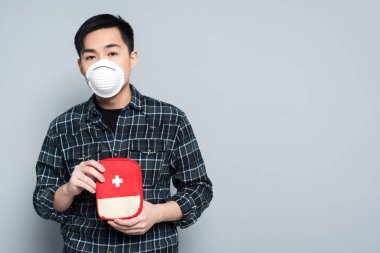 young asian man in respirator mask showing first aid kit while looking at camera isolated on grey