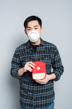 young asian man in respirator mask showing first aid kit while looking at camera on grey background
