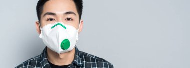 panoramic shot of young asian man in respirator mask looking at camera isolated on grey