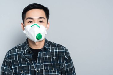 young asian man in respirator mask looking at camera while standing isolated on grey