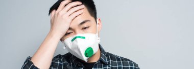panoramic shot of young asian man in respirator mask touching forehead while suffering from headache isolated on grey