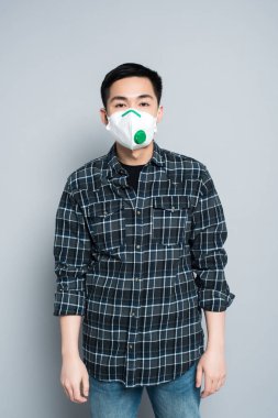 young asian man in respirator mask looking at camera while standing on grey background