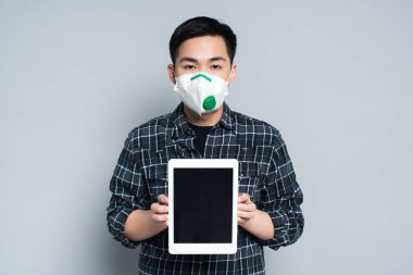 young asian man in respirator mask showing digital tablet with blank screen on grey background