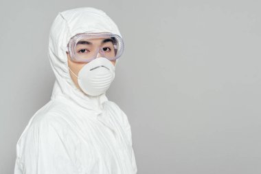 asian epidemiologist in hazmat suit and respirator mask looking at camera isolated on grey clipart