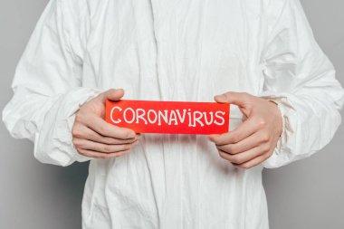 cropped view of epidemiologist in hazmat suit holding warning card with coronavirus inscription on grey background clipart
