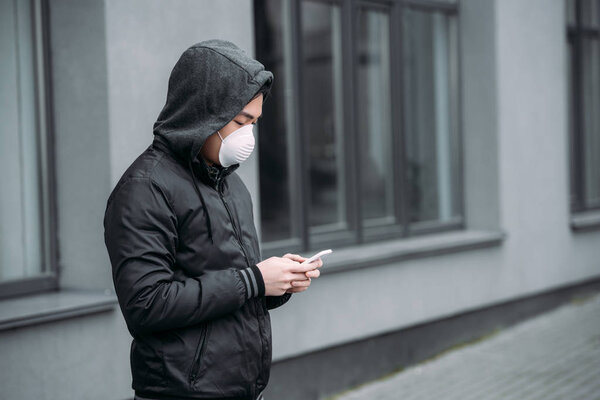 молодой азиат в респираторной маске болтает на смартфоне, стоя на улице
