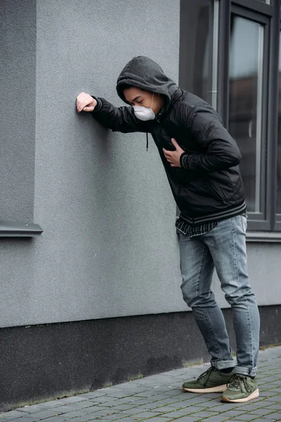 young man in respirator mask leaning on wall and touching chest while suffering from lung pain