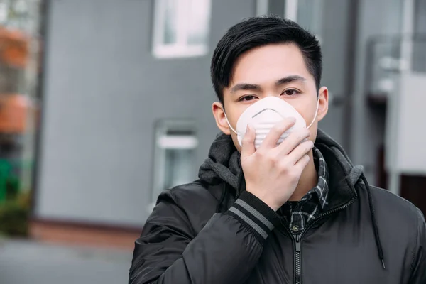 Giovane Asiatico Uomo Toccare Respiratore Maschera Guardando Fotocamera Mentre Piedi — Foto Stock