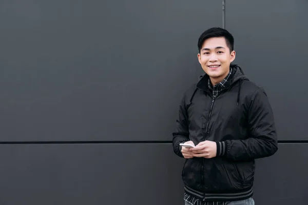 Молодой Азиат Улыбается Камеру Стоя Стены Используя Смартфон — стоковое фото