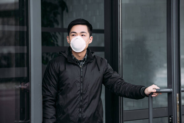 молодой азиат в респираторной маске смотрит в сторону, покидая здание

