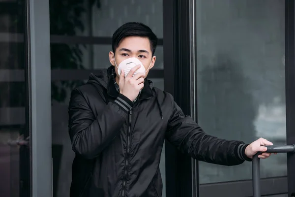 Młody Azjata Dotykając Maski Respiratora Podczas Wychodzenia Budynku Odwracając Wzrok — Zdjęcie stockowe