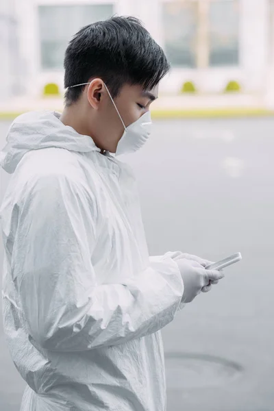 asian epidemiologist in hazmat suit and respirator mask chatting on smartphone while standing on street