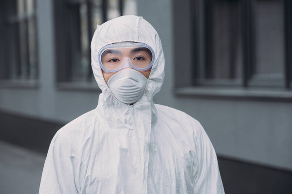 азиатский эпидемиолог в защитном костюме и респираторной маске, смотрящий в камеру, стоя рядом со зданием
