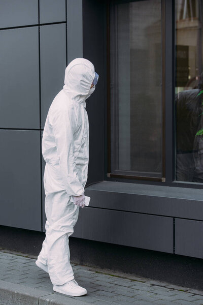эпидемиолог в защитном костюме стоит на улице и смотрит в окно здания

