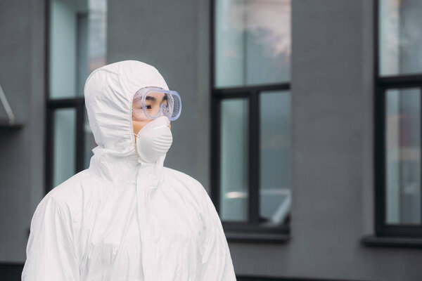 asian epidemiologist in hazmat suit and respirator mask looking away while standing near building