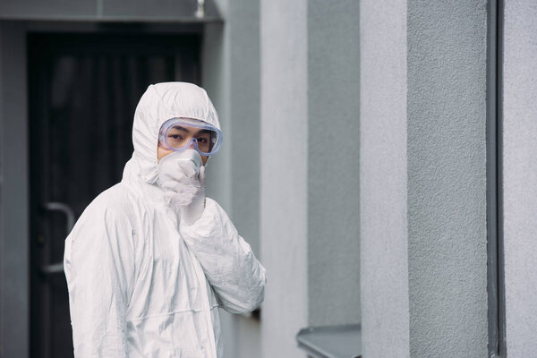 азиатский эпидемиолог в защитном костюме и респираторной маске, смотрящий в камеру, стоя снаружи

