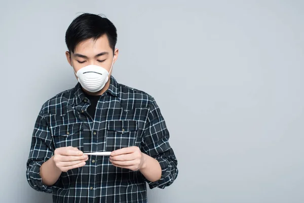 Jovem Asiático Homem Respirador Máscara Olhando Para Termômetro Cinza Fundo — Fotografia de Stock