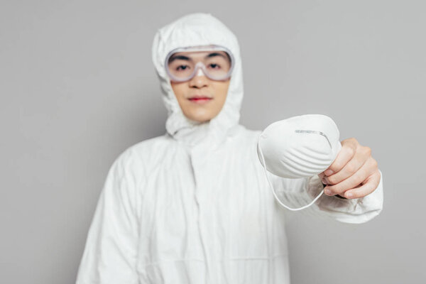 asian epidemiologist in hazmat suit showing respirator mask while looking at camera on grey background