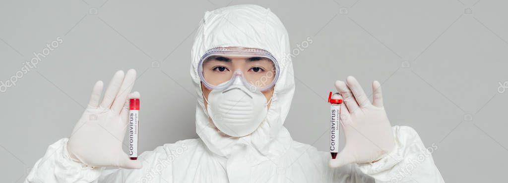 panoramic shot of asian epidemiologist in hazmat suit and respirator mask showing test tubes with blood samples isolated on grey