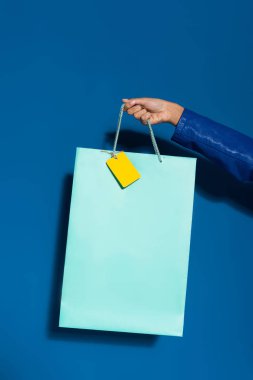 cropped view of african american woman holding shopping bag with blank label on blue