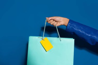 cropped view of african american woman holding shopping bag with blank label on blue