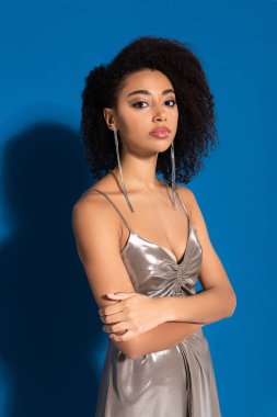 beautiful african american woman in silver dress posing with crossed arms on blue background