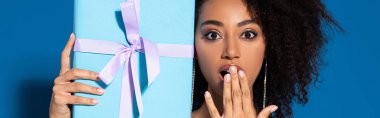 shocked beautiful african american woman holding gift and covering mouth with hand on blue background, panoramic shot