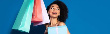 happy beautiful african american woman holding gift box and shopping bags while looking away on blue background, panoramic shot