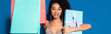 happy beautiful african american woman in silver dress holding gift box and shopping bags on blue background, panoramic shot