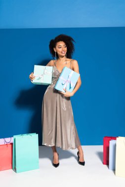 happy beautiful african american woman in silver dress holding gift boxes near shopping bags on blue background