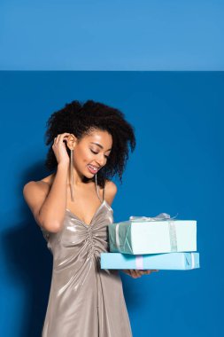 smiling beautiful african american woman in silver dress looking at gifts on blue background