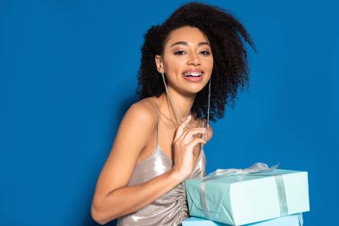smiling beautiful african american woman in silver dress holding gifts on blue background