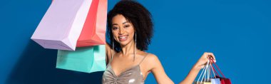 smiling elegant african american woman in silver dress holding shopping bags on blue background, panoramic shot