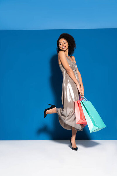 smiling beautiful african american woman in silver dress posing with shopping bags on blue background