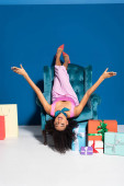usmívající se africká americká žena ležící v sametovém křesle boxu v blízkosti nákupy a dárky na modrém pozadí