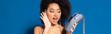 shocked beautiful african american woman in silver dress holding terminal with check on blue background, panoramic shot