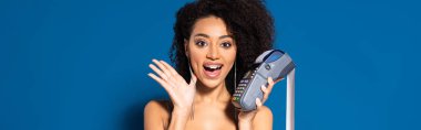 shocked beautiful african american woman in silver dress holding terminal with check on blue background, panoramic shot