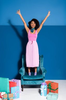 smiling african american woman standing on velour armchair with hands in air near purchases on blue background